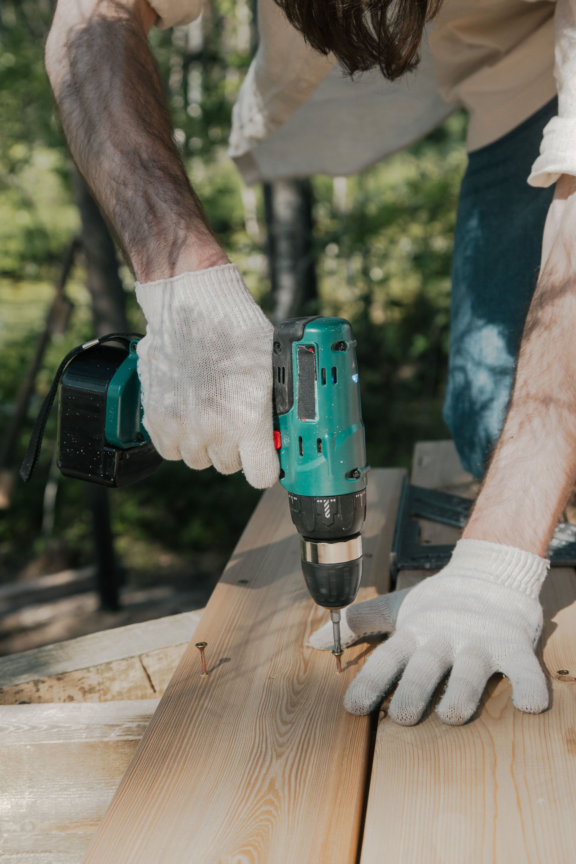 Adult using a power drill on a wooden surface, wearing white gloves, outdoors in daylight.
